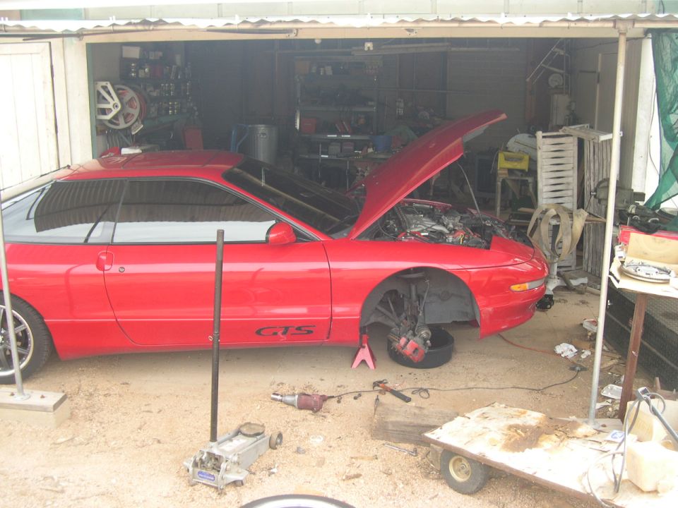 Red coupe with hood open, front wheels off, on jack stands in a garage. 'GTS' visible on door