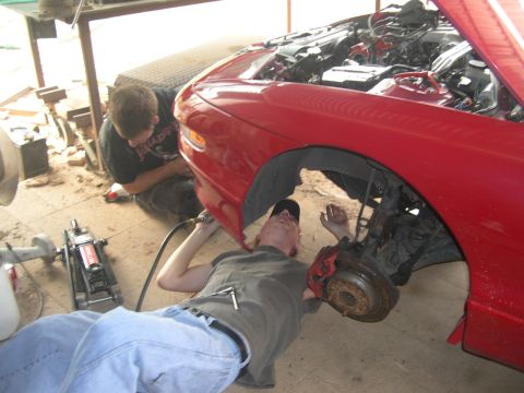 1997 Ford Probe - Two mechanics work on the front wheel assembly of a red car with its hood up.