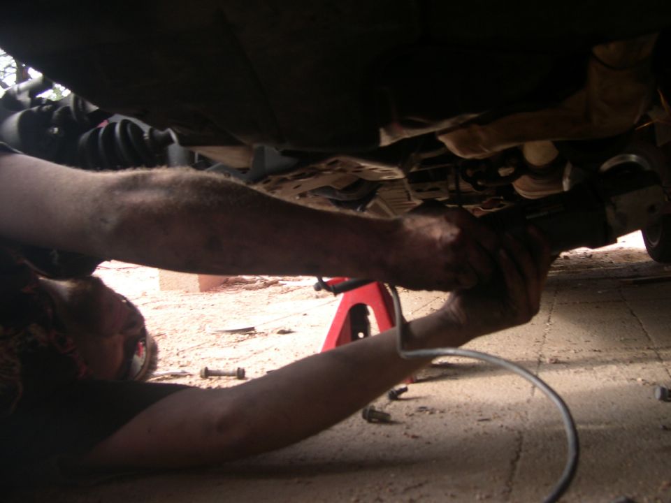 Mechanic working on car undercarriage with a tool, Supermotors.net logo visible