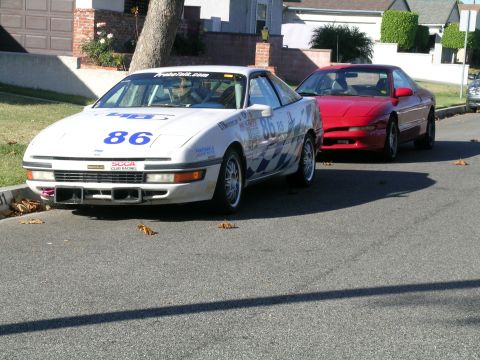 1993 Ford Probe - Two Ford Probes, one white with racing livery and the other red, are parked on a residential street.