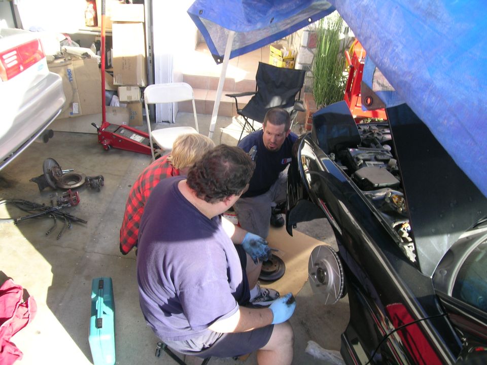 Three guys working on a black car with the hood up, one is holding a brake rotor