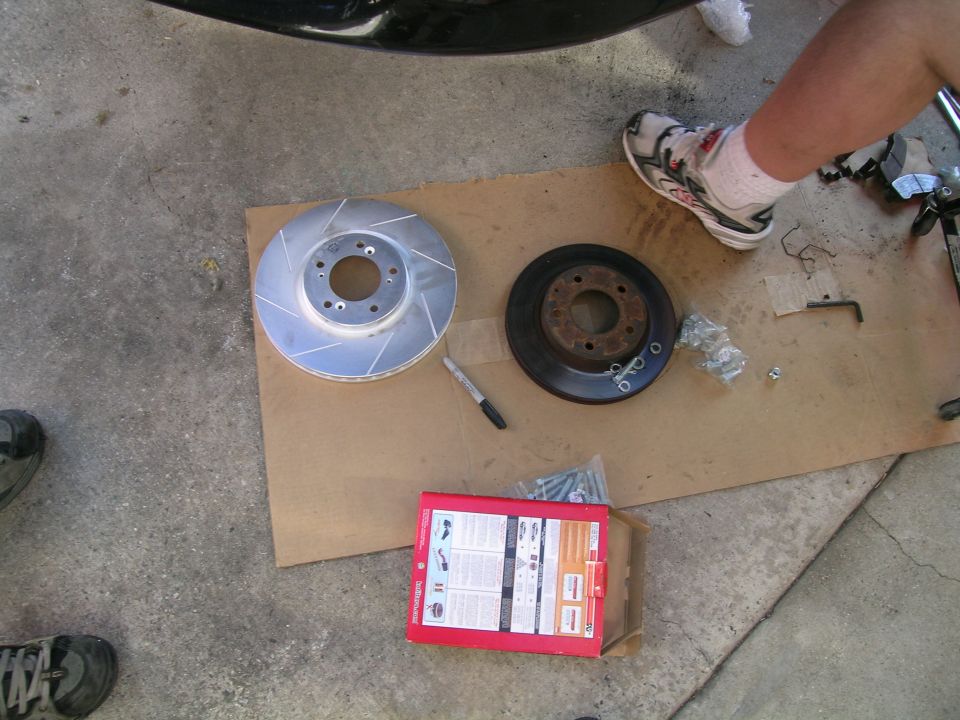 New slotted brake rotor next to old rusty one, with a box of hardware and a person's legs visible