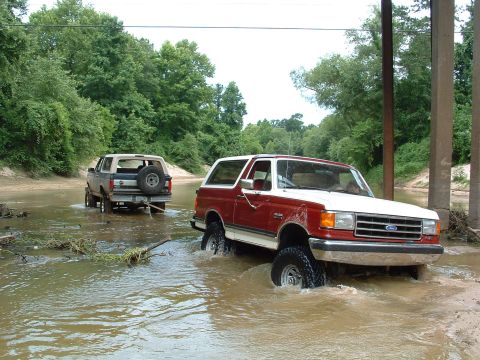 1993 Ford Bronco - Two Ford Broncos navigate a muddy, shallow waterway, kicking up water as they go.