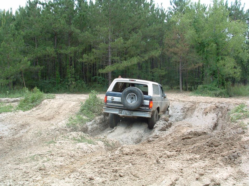 White and blue Ford Bronco driving off-road through mud, spare tire mounted on rear door