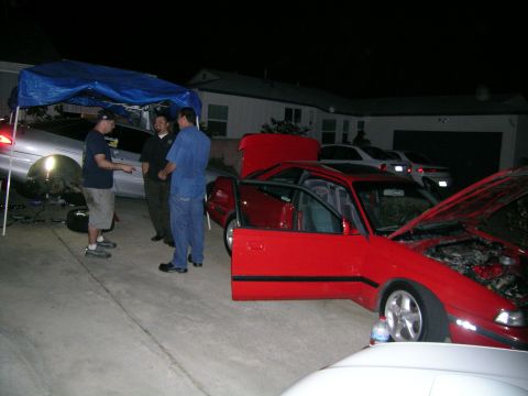 1993 Ford Probe - Several people stand around cars parked in a driveway at night, with one red coupe having its doors and hood open.