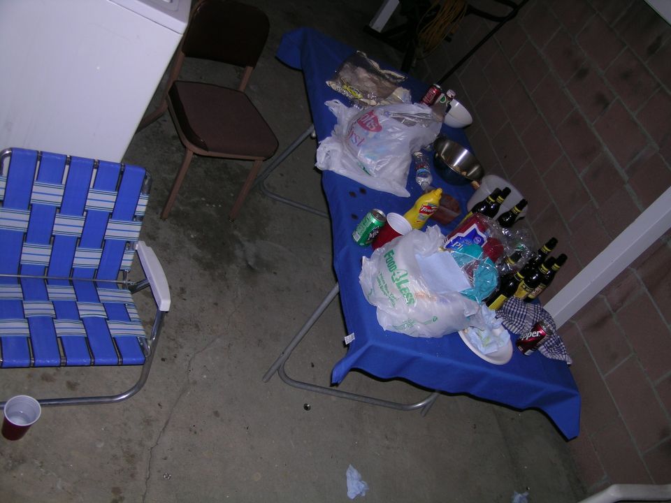 table covered in blue cloth with food, drinks, and trash, next to a lawn chair and a washing machine