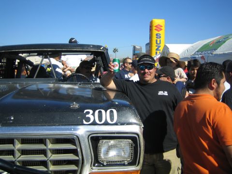 1990 Ford Bronco - man in a black shirt and sunglasses raises a drink can beside the hood of a black number 300 truck at a crowded outdoor event.