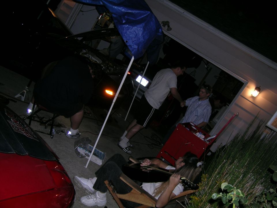 People gathered around a car in a garage, with a red car visible in the foreground