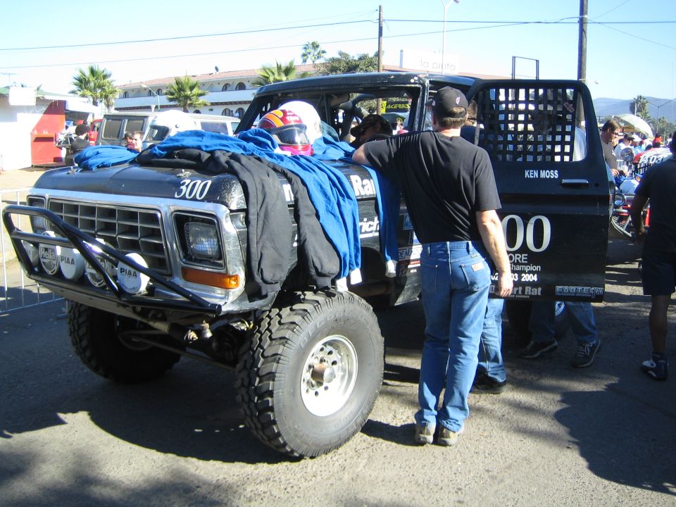 Black number 300 off-road race truck in a pit area with a man and draped tarp on hood