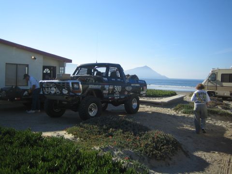 1990 Ford Bronco - black off-road race truck sits beside a house near the ocean, with a person walking nearby, an RV, and a coastal hill behind.