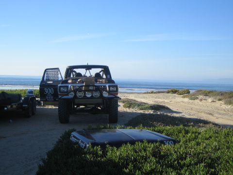 1990 Ford Bronco - black number 300 off-road race truck and trailer parked on a coastal bluff overlooking the ocean, with a 300 panel lying on the brush.