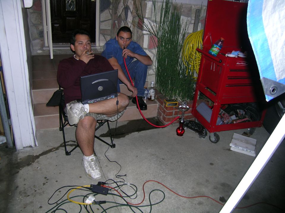 Two men sit outside with hookahs and a laptop, next to a red toolbox