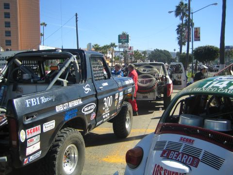 1990 Ford Bronco - black FBI Racing number 300 race truck and a white SCORE-numbered race Volkswagen Beetle staged on a palm-lined street.
