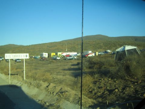 1990 Ford Bronco - dusty roadside encampment of trailers and tents seen past a sign for R El Carrizo, with hills behind.