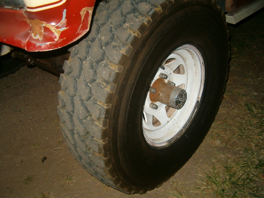 Mud-terrain tire on white spoke wheel with manual locking hub on a red vehicle, outdoors at dusk