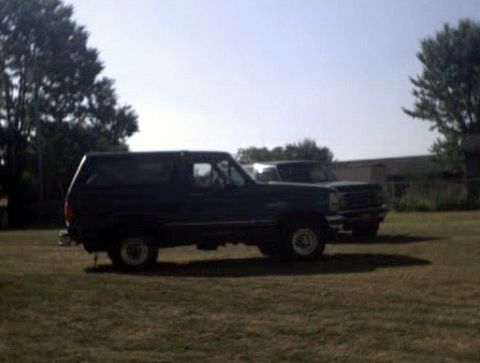 1996 Ford Bronco - A dark blue Ford Bronco is parked on a grassy field next to another vehicle under a clear sky.