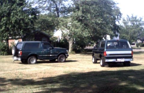 1996 Ford Bronco - Two dark green Ford Broncos are parked on a grassy area with trees and a house in the background.
