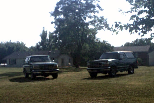 Two dark green Ford Broncos parked in a grassy yard with houses and trees in the background. The Bronco on the right is closer and more in focus