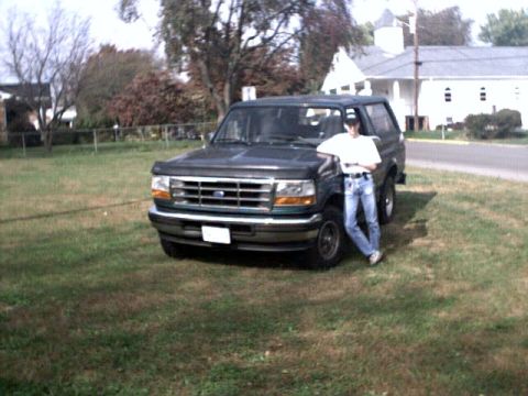 1996 Ford Bronco - A person leans against a dark green 1996 Ford Bronco parked on a grassy area next to a road.