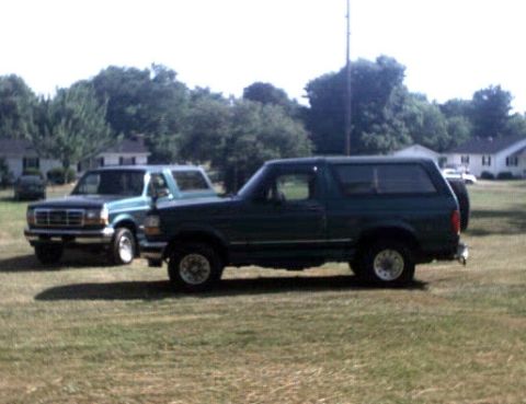 1996 Ford Bronco - Two Ford Broncos are parked in a grassy area with houses and trees in the background.