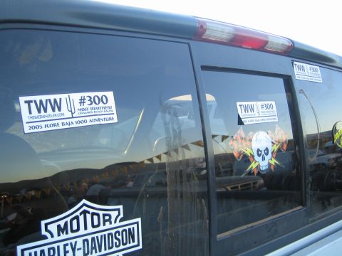 1990 Ford Bronco - side glass of a dark SUV bears two TWW #300 stickers, a flaming skull graphic and a Harley-Davidson logo.