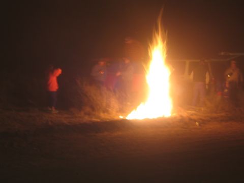 1990 Ford Bronco - tall bonfire blazes at night while several silhouetted people stand around it on bare ground.