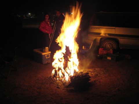 1990 Ford Bronco - bright bonfire burns at night next to a light van, with two people and camp coolers beside it.