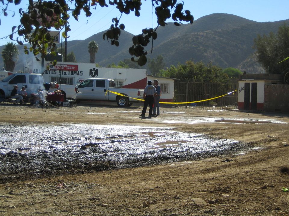 Muddy dirt lot with white vans, people behind yellow caution tape and hills under a clear sky