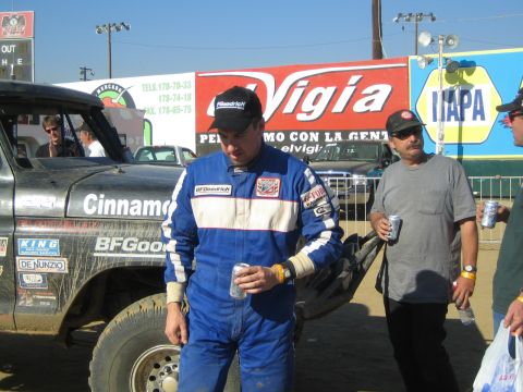1990 Ford Bronco - man in a blue BFGoodrich racing suit holds a can beside a black Cinnamon race truck under El Vigia signage.
