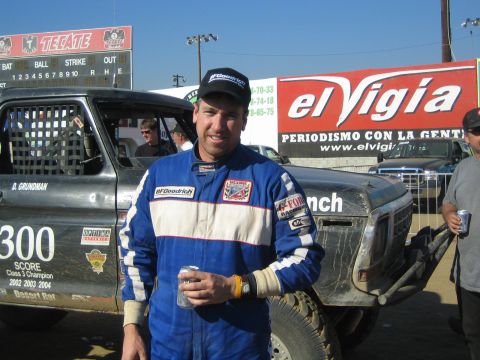 1990 Ford Bronco - smiling man in a blue BFGoodrich race suit and cap holds a can next to a black 300 SCORE race truck.