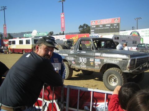 1990 Ford Bronco - man in a cap leans on a barrier as a black Cinnamon Ranch 300 race truck sits behind, baseball scoreboard above.