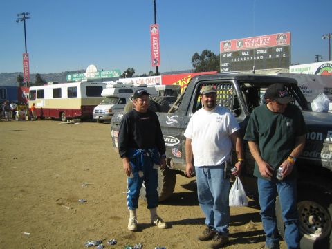 1990 Ford Bronco - Three men holding drinks stand on a dirt lot beside off-road trucks and RVs below a Tecate baseball scoreboard.