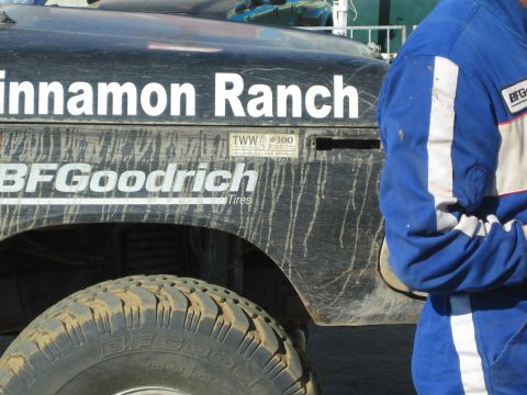 1990 Ford Bronco - Close-up of a mud-streaked black race truck panel lettered Cinnamon Ranch and BFGoodrich Tires beside a person in blue.