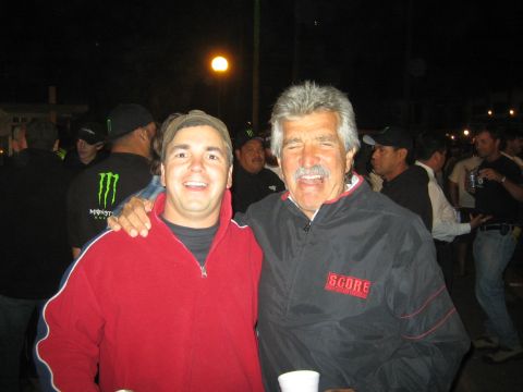 1990 Ford Bronco - young man in a red hoodie poses arm-in-arm with a smiling older mustached man wearing a black SCORE jacket at a crowded night event.