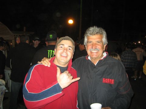 1990 Ford Bronco - young man in a red zip-up throws a shaka sign while posing with a smiling older mustached man in a black SCORE jacket at a night event.