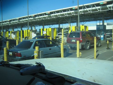 1990 Ford Bronco - line of cars and a maroon SUV wait at lanes of a border crossing flanked by yellow bollards and an overhead steel canopy.