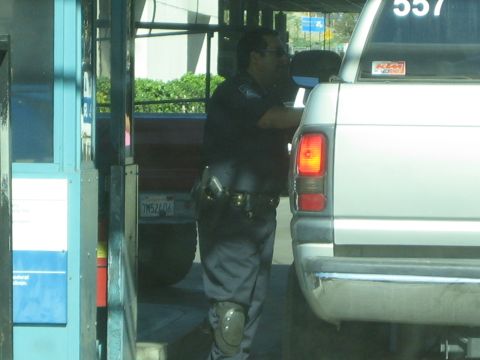 1990 Ford Bronco - uniformed officer with a duty belt stands beside the rear of a silver pickup near a crossing booth, seen through a window.