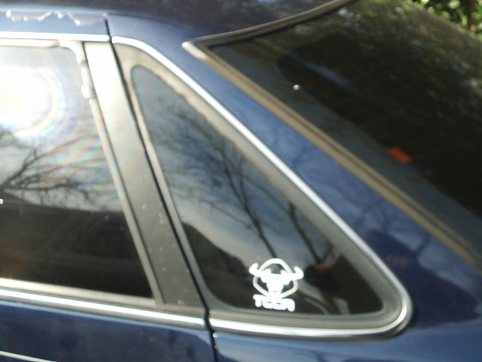 Dark blue car's rear side window with a white sticker of a bull's head and the word 'TOEA' below it. Supermotors.net logo in lower right