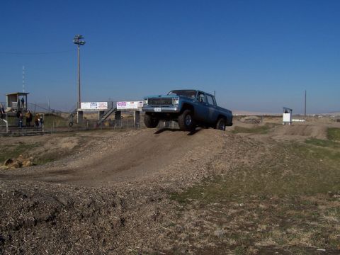 1979 Ford Bronco - A blue GMC pickup truck navigates an off-road course, climbing a dirt ramp under a clear blue sky.
