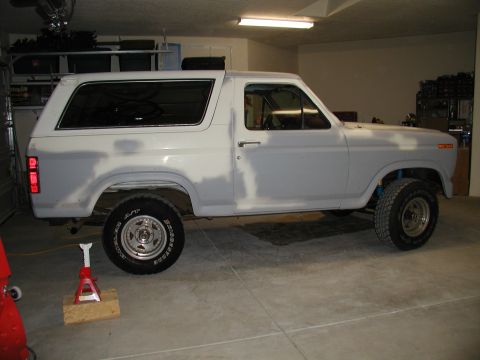 1986 Ford Bronco - A Ford Bronco is parked in a garage, showing primer on its body panels and a jack supporting the rear passenger side wheel.