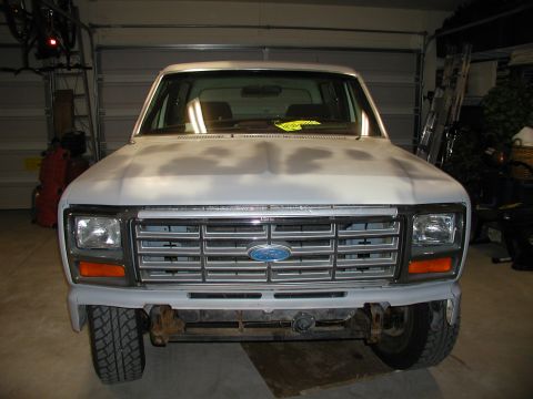 1986 Ford Bronco - Front view of a white Ford Bronco with a primer hood and chrome grille, parked indoors.