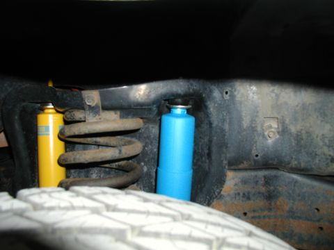 1986 Ford Bronco - Close-up of the undercarriage shows a rusty coil spring and two shock absorbers, one yellow and one blue.