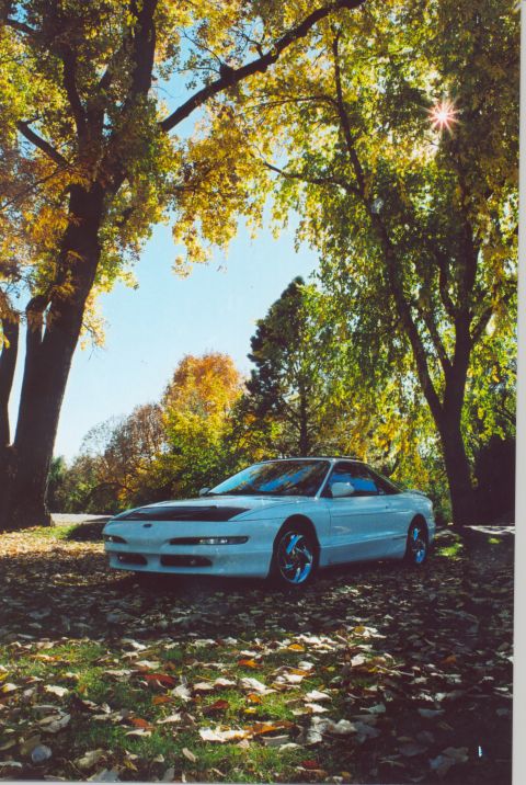 1996 Ford Probe - A white coupe sits parked on the ground covered in fallen leaves, surrounded by trees in autumn.