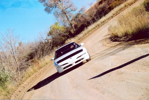 1996 Ford Probe - A white sports coupe navigates a dusty, winding dirt road under a clear blue sky.
