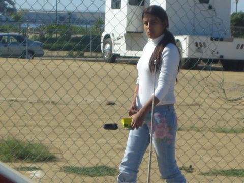 1990 Ford Bronco - woman in a white turtleneck and floral-print jeans stands behind a chain-link fence with a parked trailer and dirt lot beyond.