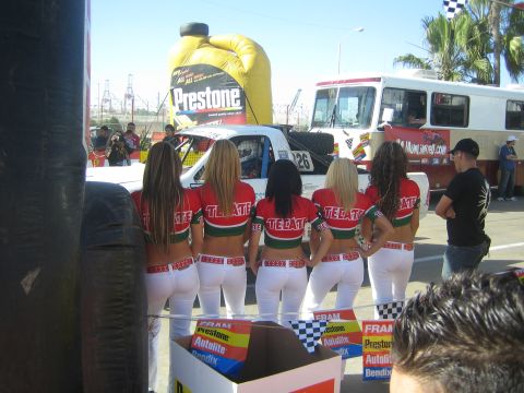 1990 Ford Bronco - Five promo models in red Tecate tops and white pants stand in a row facing a race car and bus, with Prestone and FRAM signage nearby.