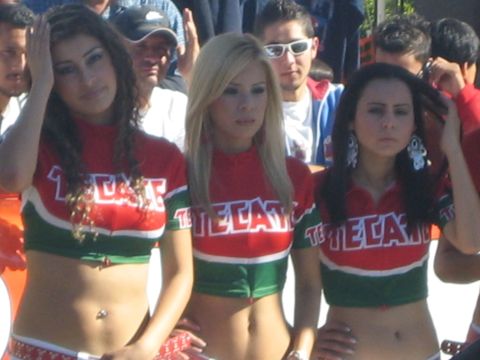 1990 Ford Bronco - Three promo models in red and green Tecate crop tops pose for the camera with a crowd of spectators behind them at an outdoor event.