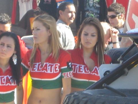 1990 Ford Bronco - Promo models in red and green Tecate crop tops stand among spectators beside a large mud-terrain tire at an outdoor event.