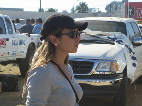 1990 Ford Bronco - woman in a black cap and sunglasses stands in profile beside a white off-road pickup marked Baja 1000 in a dirt staging area.
