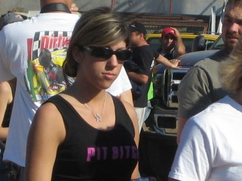 1990 Ford Bronco - woman in sunglasses wears a black tank top reading PIT BITCH amid a crowd, with trucks and a graphic T-shirt visible behind her.